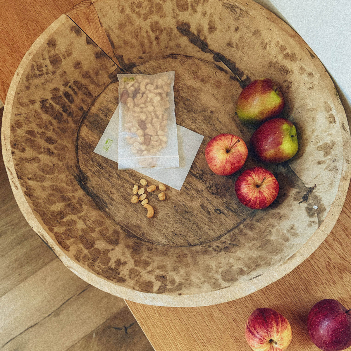 Top view of a rustic, wooden bowl filled with a transparent bag of mixed nuts, some individual nuts and three red apples.