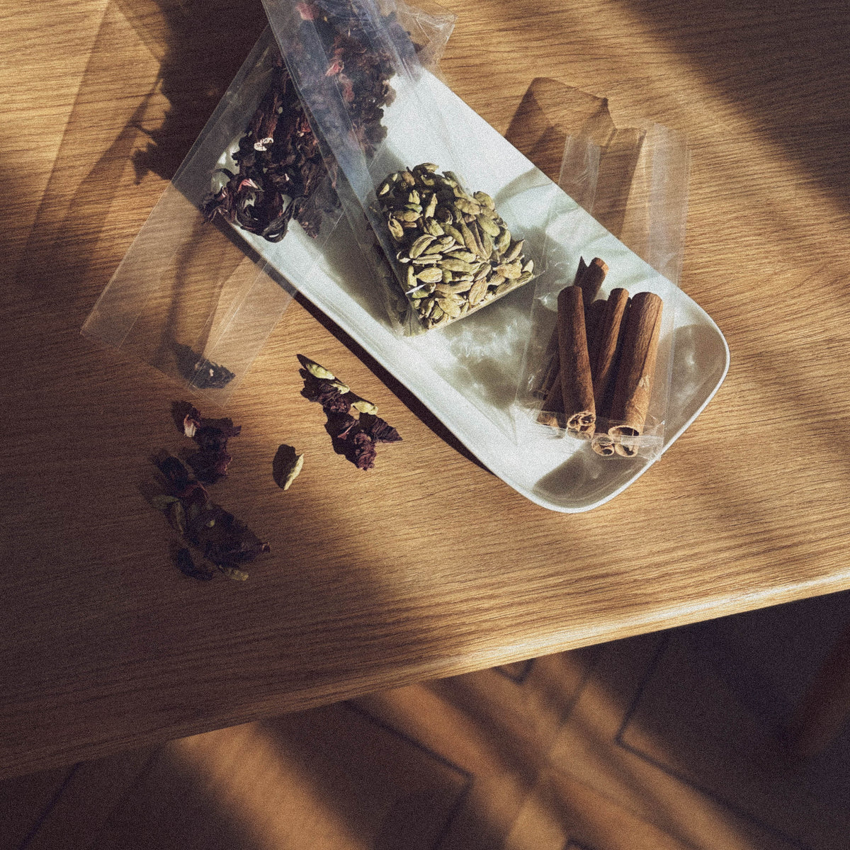 Top view of an elongated, white bowl standing on a light-colored wooden surface and containing various spices in transparent plastic bags: dried hibiscus flowers, green cardamom pods and cinnamon sticks.