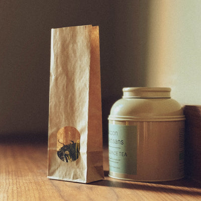 A narrow, brown paper bag with a round sticker and a round, light green metal tin with a label, standing on a wooden surface against a dark background.